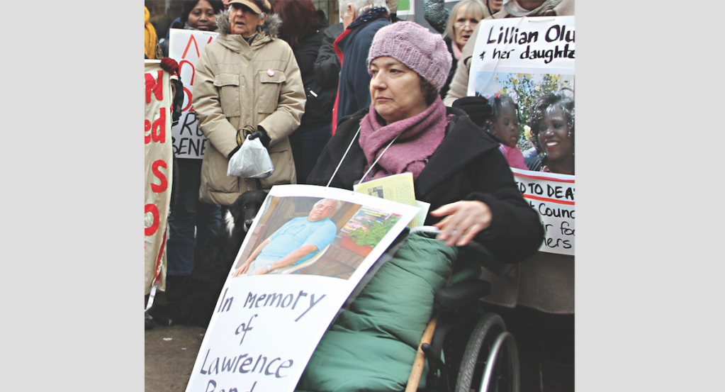 Close-up photo from a vigil in January 2017 at Kentish Town Jobcentre in memory of Lawrence Bond.  A white woman wheelchair user holds a placard with a photo of Lawrence Bond.  Behind her, a woman of colour holds a placard in memory of Lillian Oluk and her baby daughter who starved to death. Behind them various people on the vigil, some holding banners and placards.